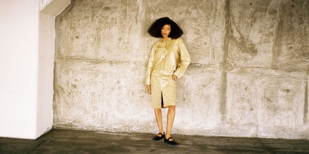 kilo-kish-wears-a-tan-skirt-and-button-up-shirt-and-stands-at-a-distance-leaning-against-a-wall.jpg