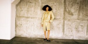 kilo-kish-wears-a-tan-skirt-and-button-up-shirt-and-stands-at-a-distance-leaning-against-a-wall.jpg
