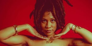 a-black-woman-with-dreadlocks-and-gold-jewlery-poses-with-her-hands-under-her-chin-against-a-red-background.jpg