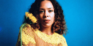 a-woman-with-a-yellow-shirt-holds-a-yellow-flower-against-a-blue-background.png