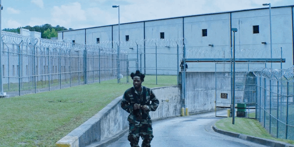 shameik-moore-wearing-an-all-camo-outfit-and-leaving-what-appears-to-be-a-prison.png