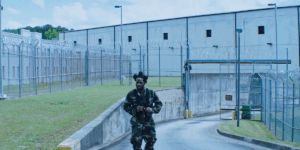 shameik-moore-wearing-an-all-camo-outfit-and-leaving-what-appears-to-be-a-prison.png