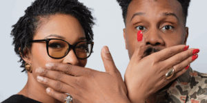 two-people-stand-posing-with-their-hands-playfully-covering-each-other-s-mouth.png